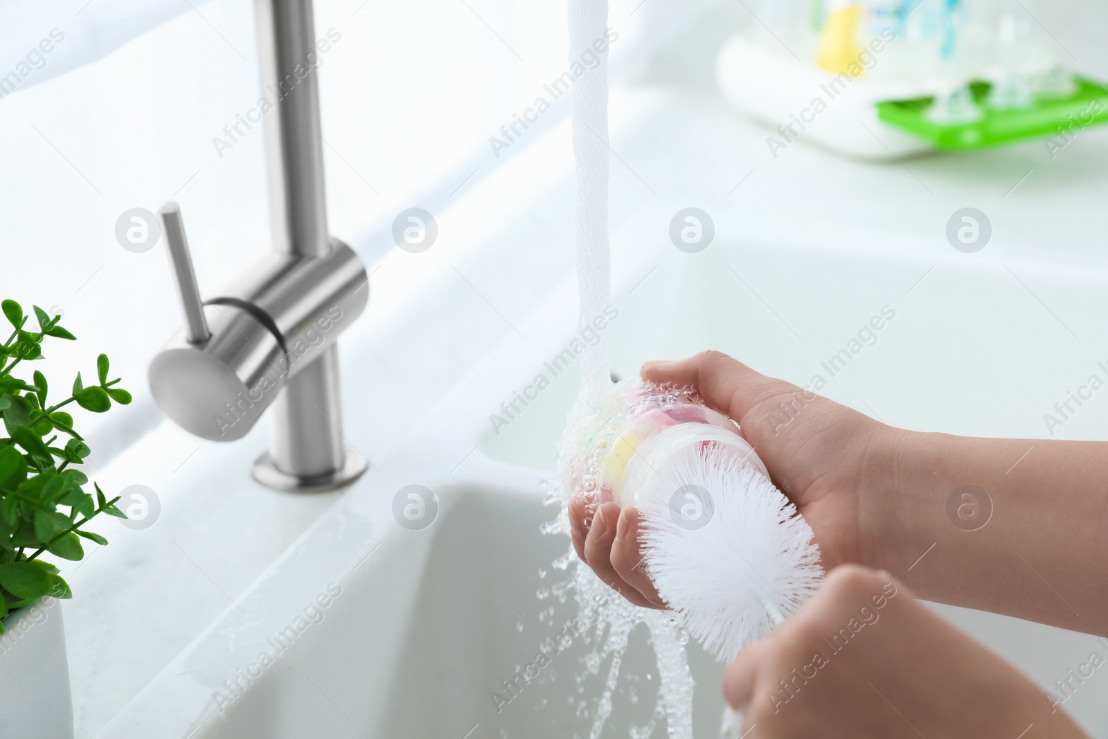 Photo of Woman washing baby bottle under stream of water in kitchen, closeup