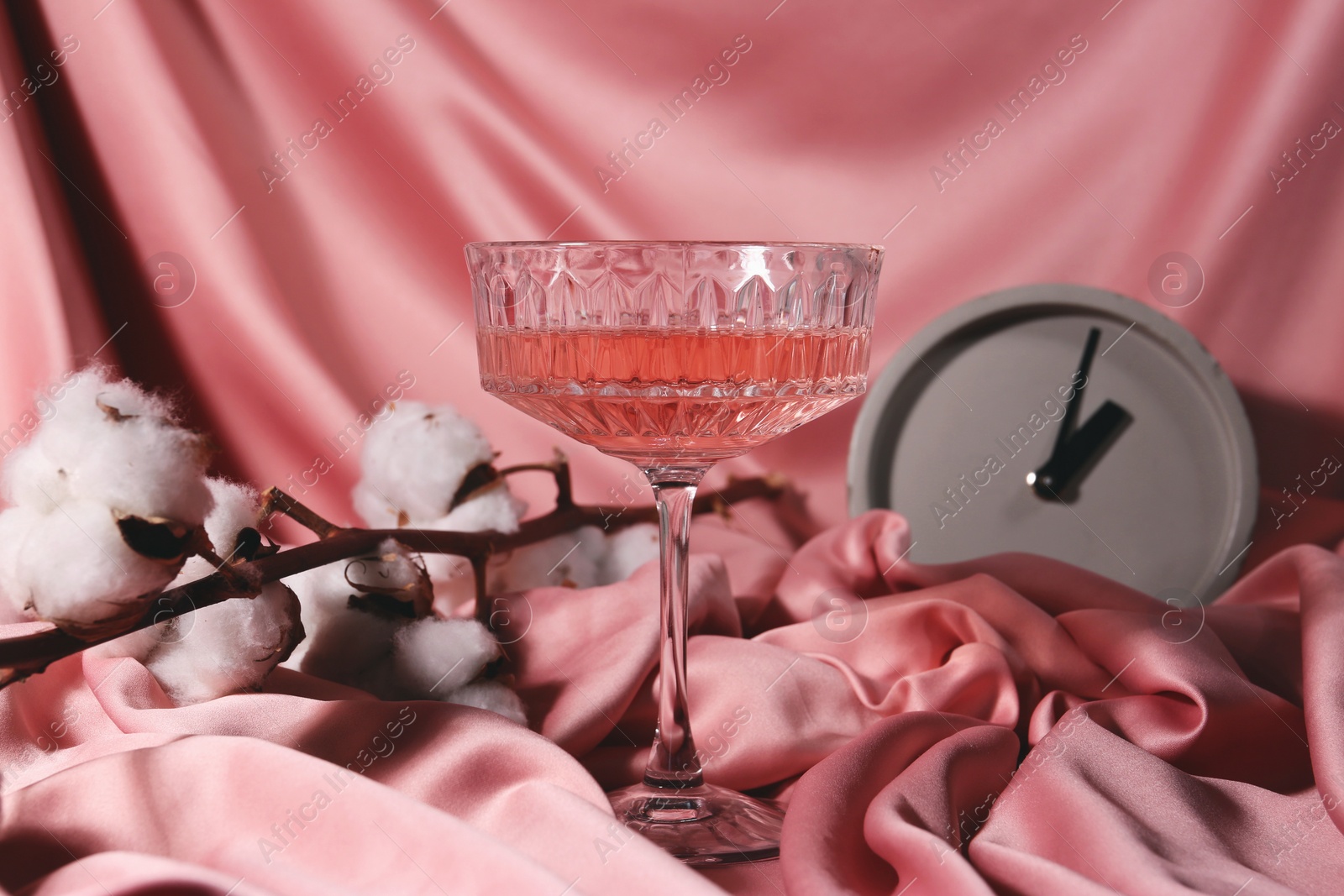 Photo of Glass of alcohol drink, clock and cotton flowers on pink cloth