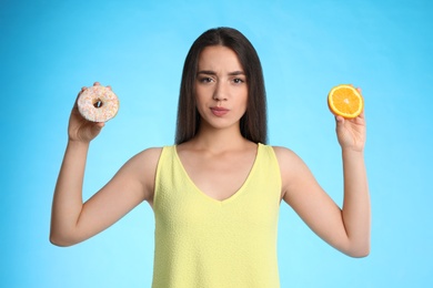 Photo of Doubtful woman choosing between orange and doughnut on light blue background