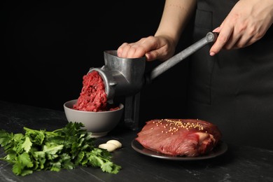 Woman making beef mince with manual meat grinder at dark textured table against black background, closeup