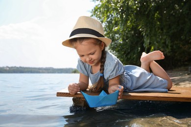 Cute little girl playing with paper boat on wooden pier near river