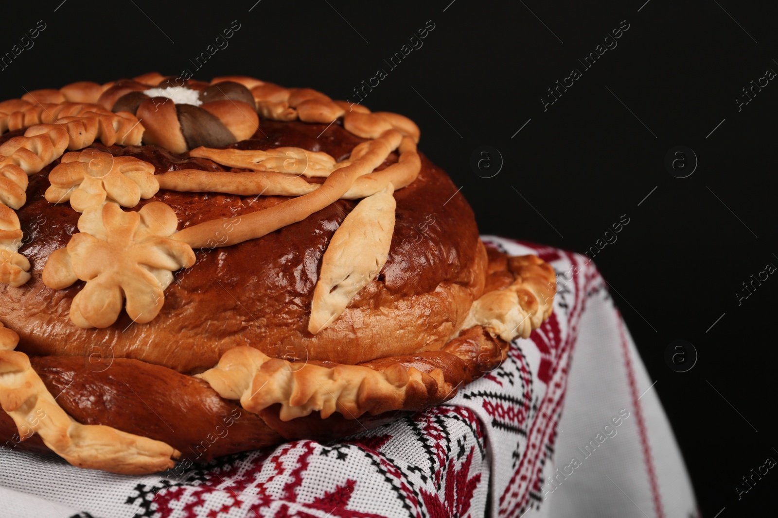 Photo of Rushnyk with korovai on table against black background, closeup. Ukrainian bread and salt welcoming tradition