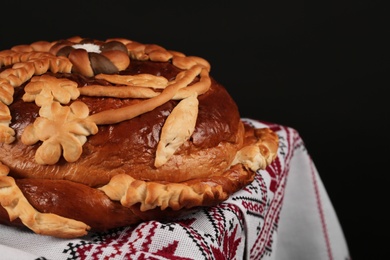 Photo of Rushnyk with korovai on table against black background, closeup. Ukrainian bread and salt welcoming tradition