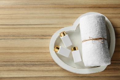 Top view of heart shaped tray with towel and cosmetic bottles on wooden table, space for text. Spa treatment