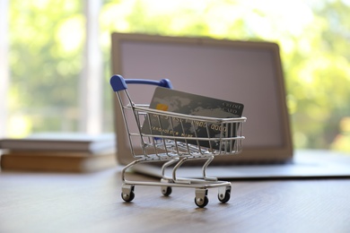 Photo of Internet shopping. Small cart with credit card near laptop on table indoors