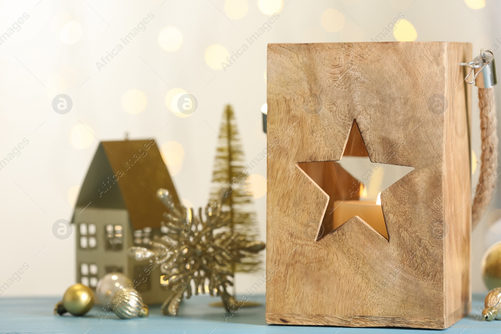 Photo of Lantern with burning candle and Christmas decorations on light blue wooden table against blurred lights. Space for text