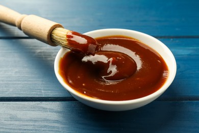 Tasty barbeque sauce in bowl and brush on blue wooden table, closeup