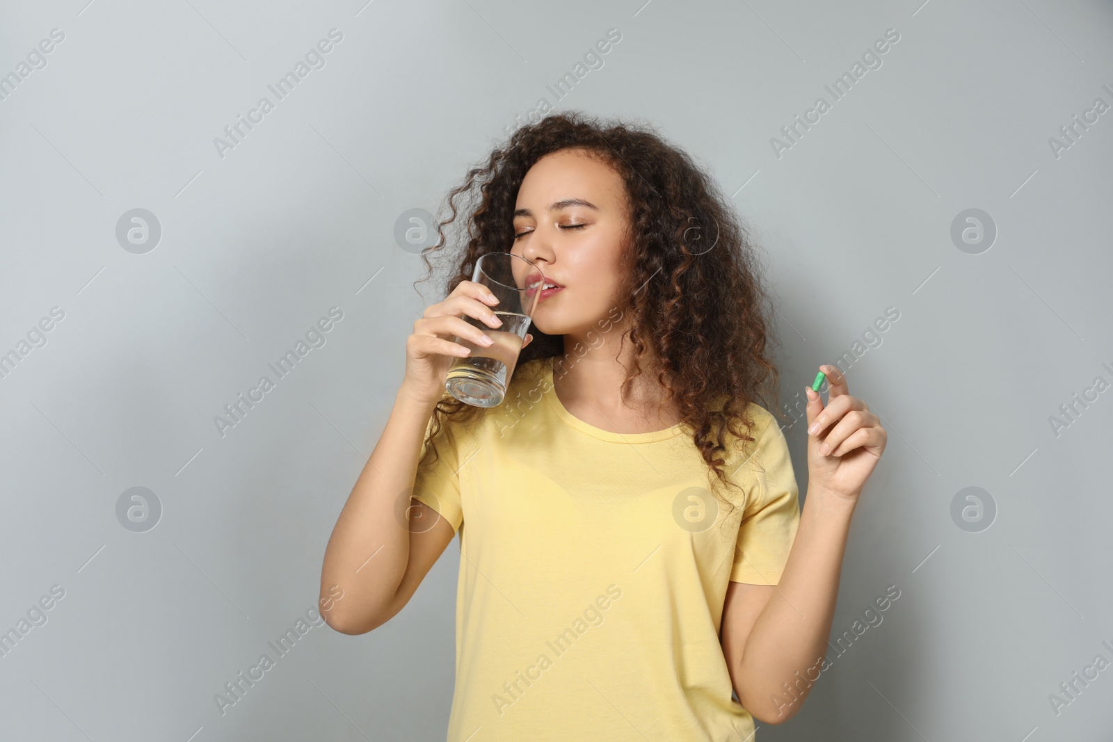 Photo of African-American woman with glass of water and vitamin capsule on light grey background