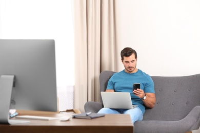 Handsome young man working with smartphone and laptop on sofa indoors