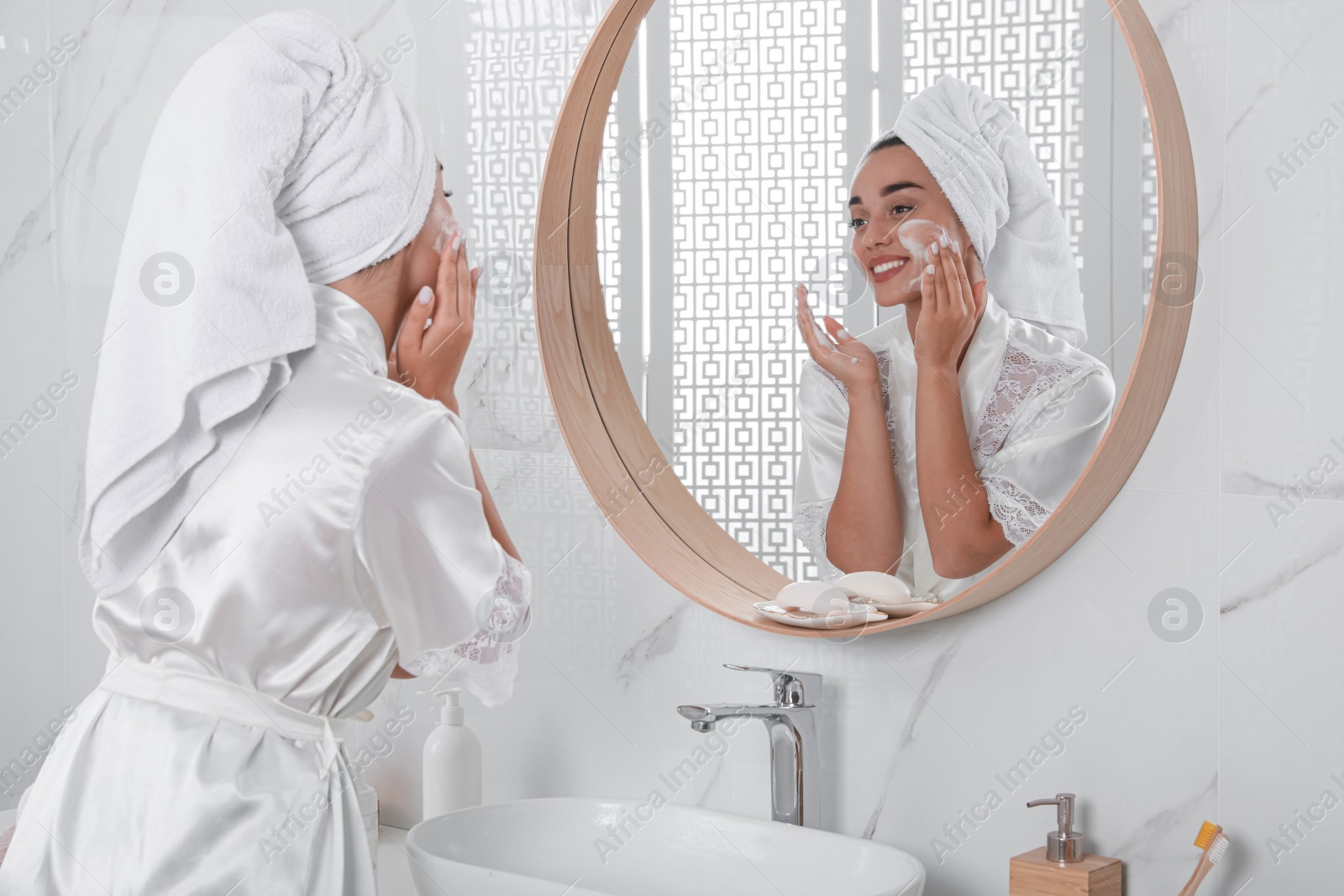 Photo of Beautiful young woman applying cleansing foam onto face near mirror in bathroom. Skin care cosmetic