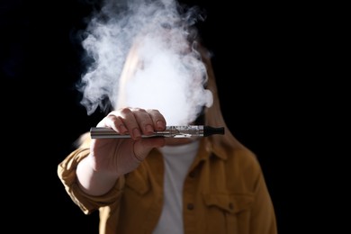 Young woman holding electronic cigarette against black background, focus on hand