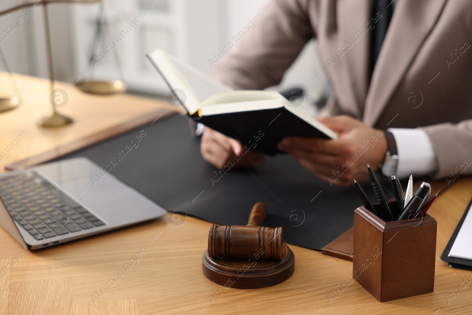 Photo of Lawyer reading book at table in office, focus on gavel and pens holder