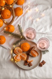 Photo of Delicious ripe tangerines, chocolates, festive lights and glasses of wine on white bedsheet, flat lay