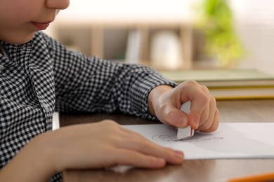 Little boy erasing mistake in his notebook at wooden desk indoors, closeup