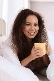 Happy African American woman with cup of drink in bed at home