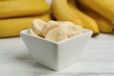 Photo of Bowl with cut bananas near whole fruits on white wooden table, closeup