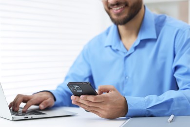 Man using smartphone while working with laptop at white table in office, closeup