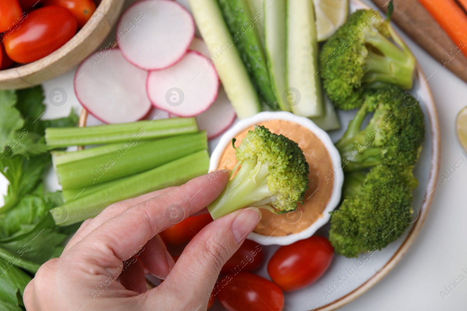 Photo of Woman dipping broccoli into hummus at white table, top view