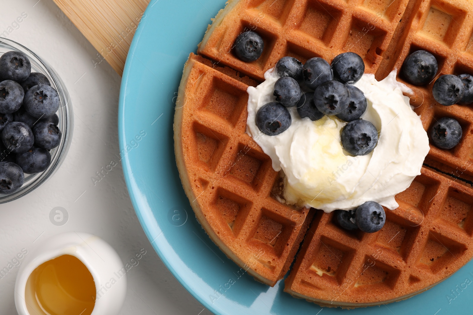 Photo of Tasty Belgian waffle with blueberries, honey and whipped cream on white table, flat lay