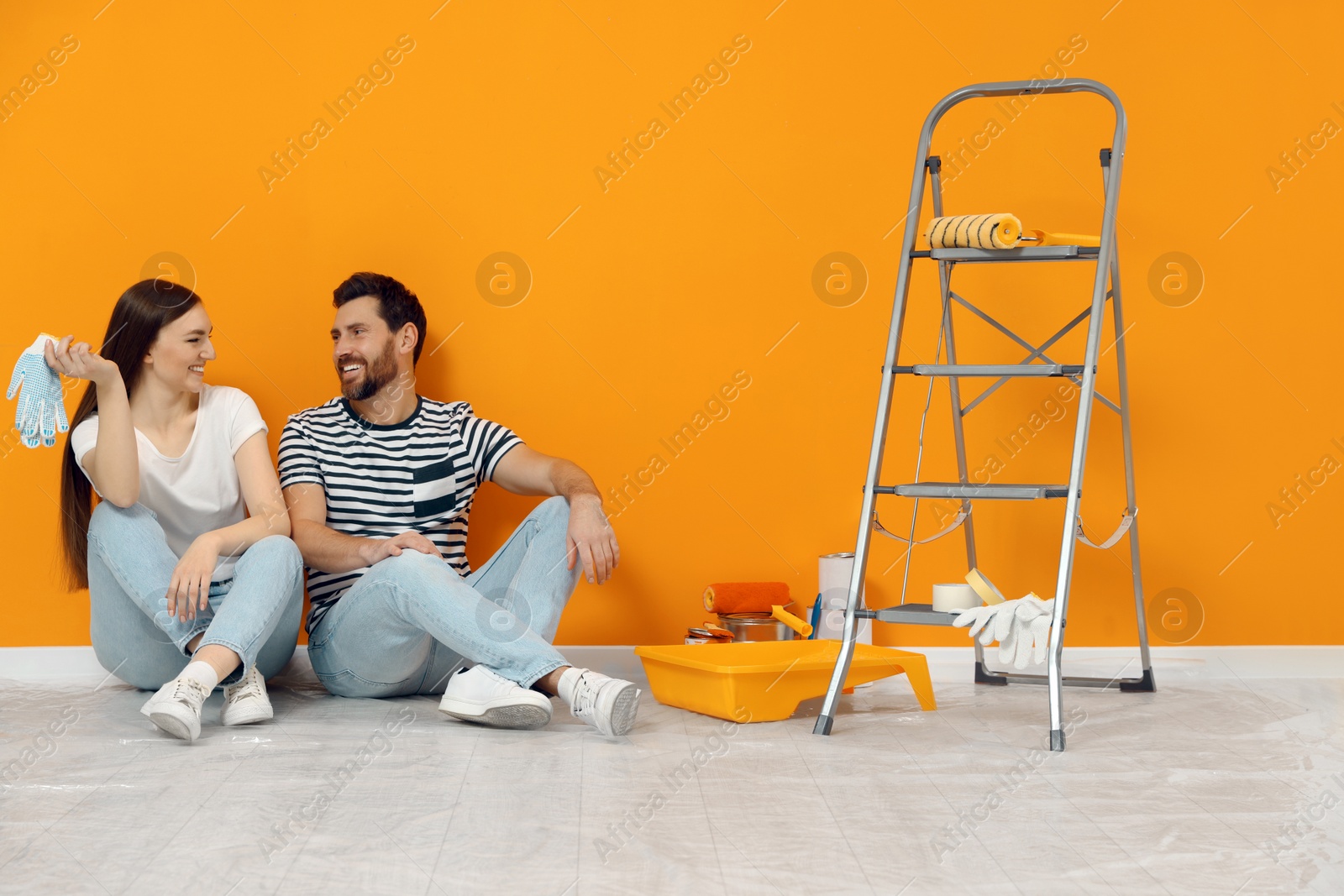 Photo of Happy designers sitting on floor with painting equipment near freshly painted orange wall indoors