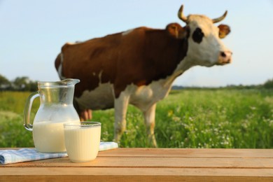 Photo of Glass with jug of milk on wooden table and cow grazing in meadow