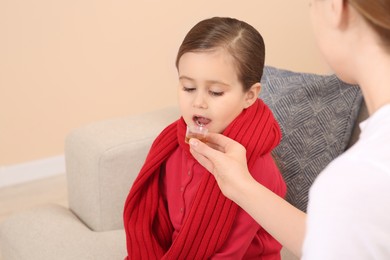 Mother giving cough syrup to her daughter from measuring cup on sofa indoors