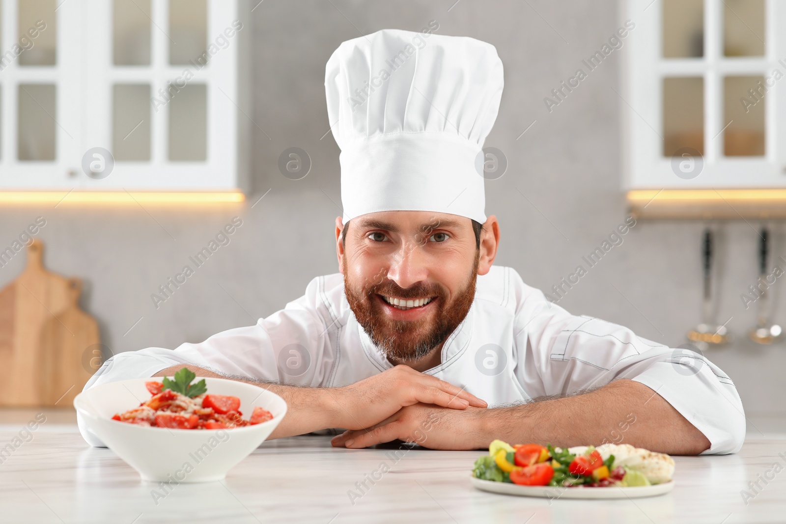 Photo of Happy professional chef near delicious food at marble table in kitchen