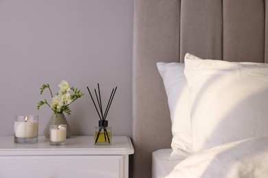 Photo of Aromatic reed air freshener, freesia flowers and candles on white bedside table in bedroom