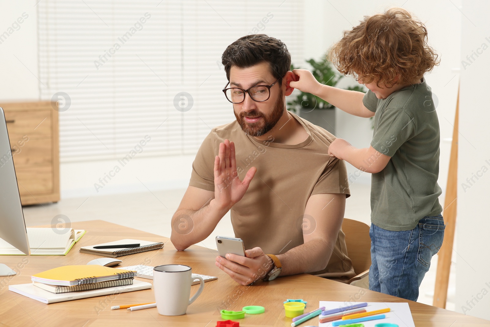 Photo of Little boy bothering his father at home. Man working remotely at desk