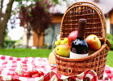 Photo of Picnic basket with fruits and bottle of wine on checkered blanket in garden