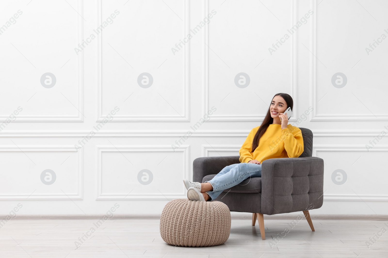 Photo of Beautiful woman talking on smartphone while sitting in armchair near white wall indoors, space for text