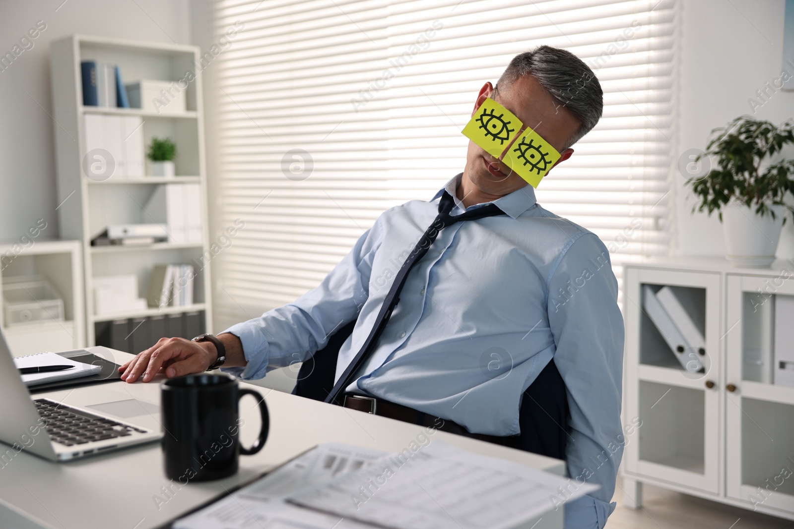 Photo of Man with fake eyes painted on sticky notes snoozing at workplace in office