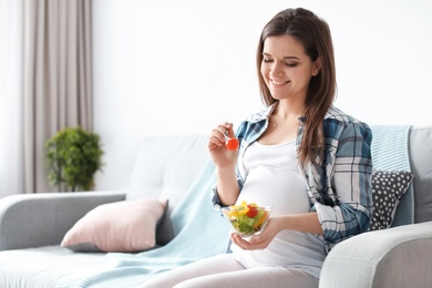 Photo of Young pregnant woman eating vegetable salad on sofa at home