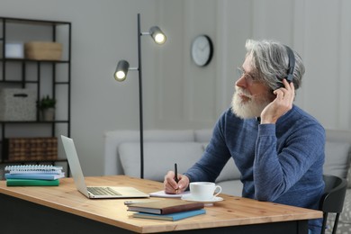 Photo of Middle aged man with laptop, notebook and headphones learning at table indoors