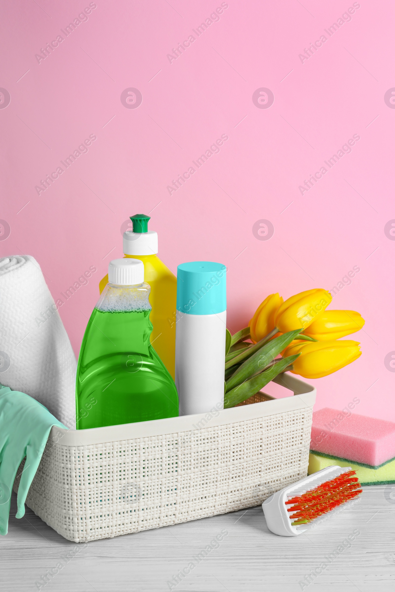 Photo of Plastic basket with different cleaning supplies and beautiful spring flowers on white wooden table against light pink background