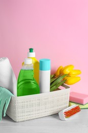 Plastic basket with different cleaning supplies and beautiful spring flowers on white wooden table against light pink background