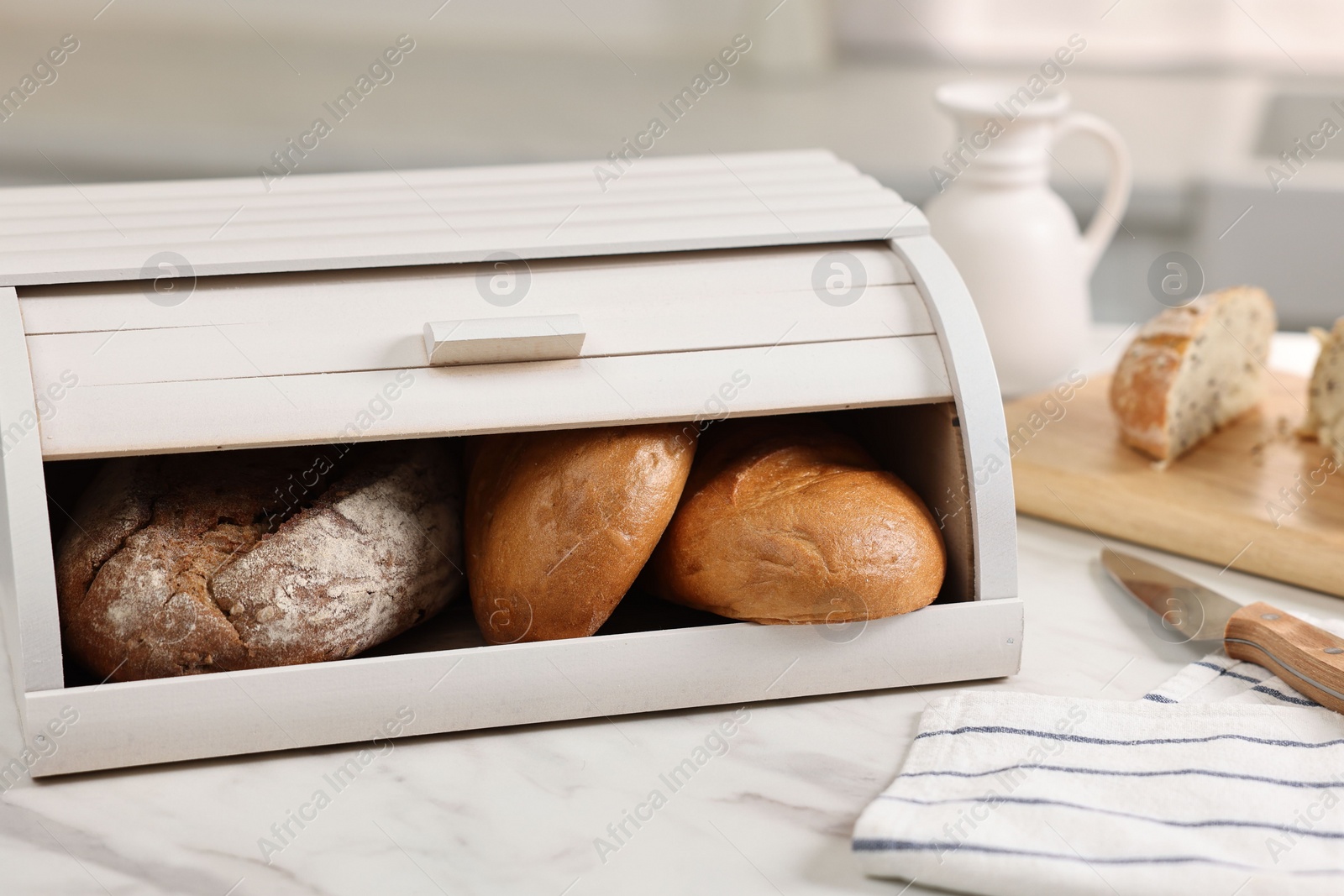 Photo of Wooden bread basket with freshly baked loaves and knife on white marble table in kitchen