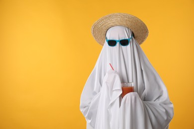 Photo of Person in ghost costume, sunglasses and straw hat holding glass of drink on yellow background, space for text