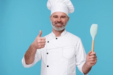 Photo of Happy chef in uniform with spatula showing thumbs up on light blue background