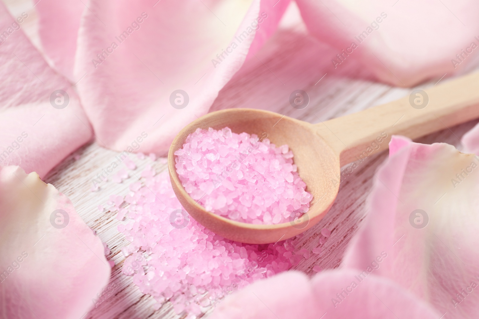 Photo of Spoon with pink sea salt and petals of roses on white wooden table, closeup