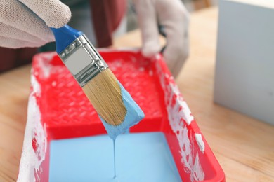 Woman taking light blue paint with brush from tray at wooden table, closeup