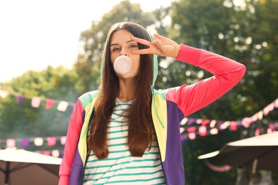 Beautiful young woman blowing chewing gum and showing peace gesture outdoors