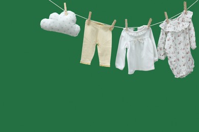 Different baby clothes and cloud shaped pillow drying on laundry line against green background. Space for text