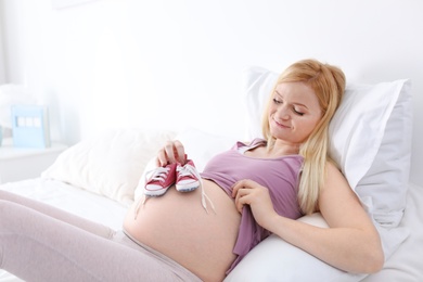 Photo of Beautiful pregnant woman holding baby booties near tummy on bed in light room