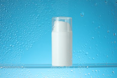 Photo of Bottle with moisturizing cream on light blue background, view through wet glass