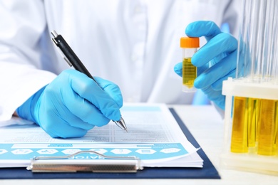 Laboratory assistant with urine sample for analysis writing medical report at table, closeup