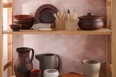 Photo of Set of different crafting tools and clay dishes on wooden rack in workshop