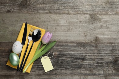 Photo of Cutlery set, painted eggs and beautiful flower on wooden table, flat lay with space for text. Easter celebration