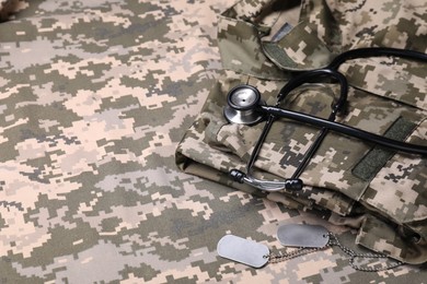 Photo of Stethoscope, military ID tags and soldier uniform on camouflage fabric. Space for text