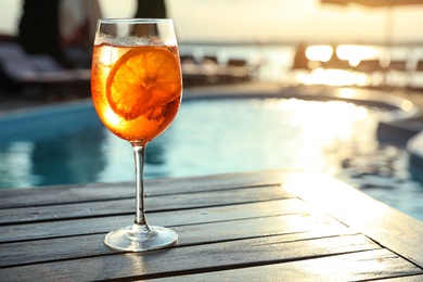 Glass of fresh summer cocktail on wooden table near swimming pool outdoors at sunset. Space for text